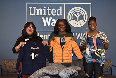Three women each stand and holding a coat in front of the United Way logo during the coat drive