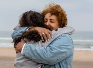 two women hugging on a beach. One is wearing a jean jacket the other a grey sweatshirt. It looks like a cold and cloudy day.
