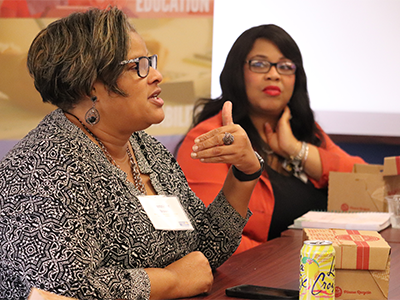 Image of an AALC member talking at a meeting with her hands. She is wearing a black pattern jacket and black glasses