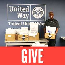Click here to give - Man holding a box filled with bags of books in front of a Trident United Way sign