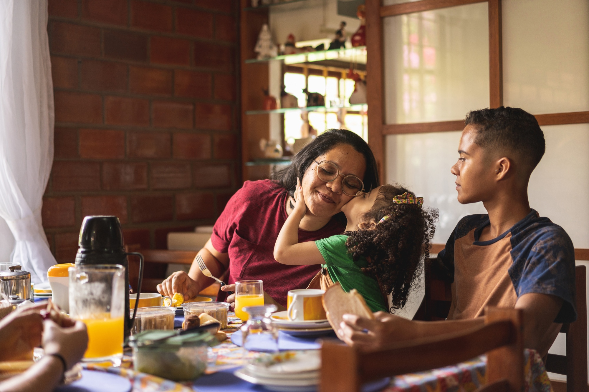 A young boy, young girl and older woman sit around the table eating. The young girl is kissing the older woman on the cheek and the younger boy looks on.