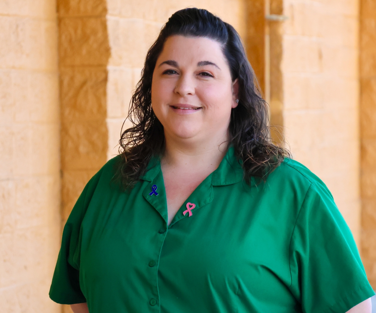 Photo of a woman standing outside in front of a yellow building wearing a green shirt and smiling at the camera.