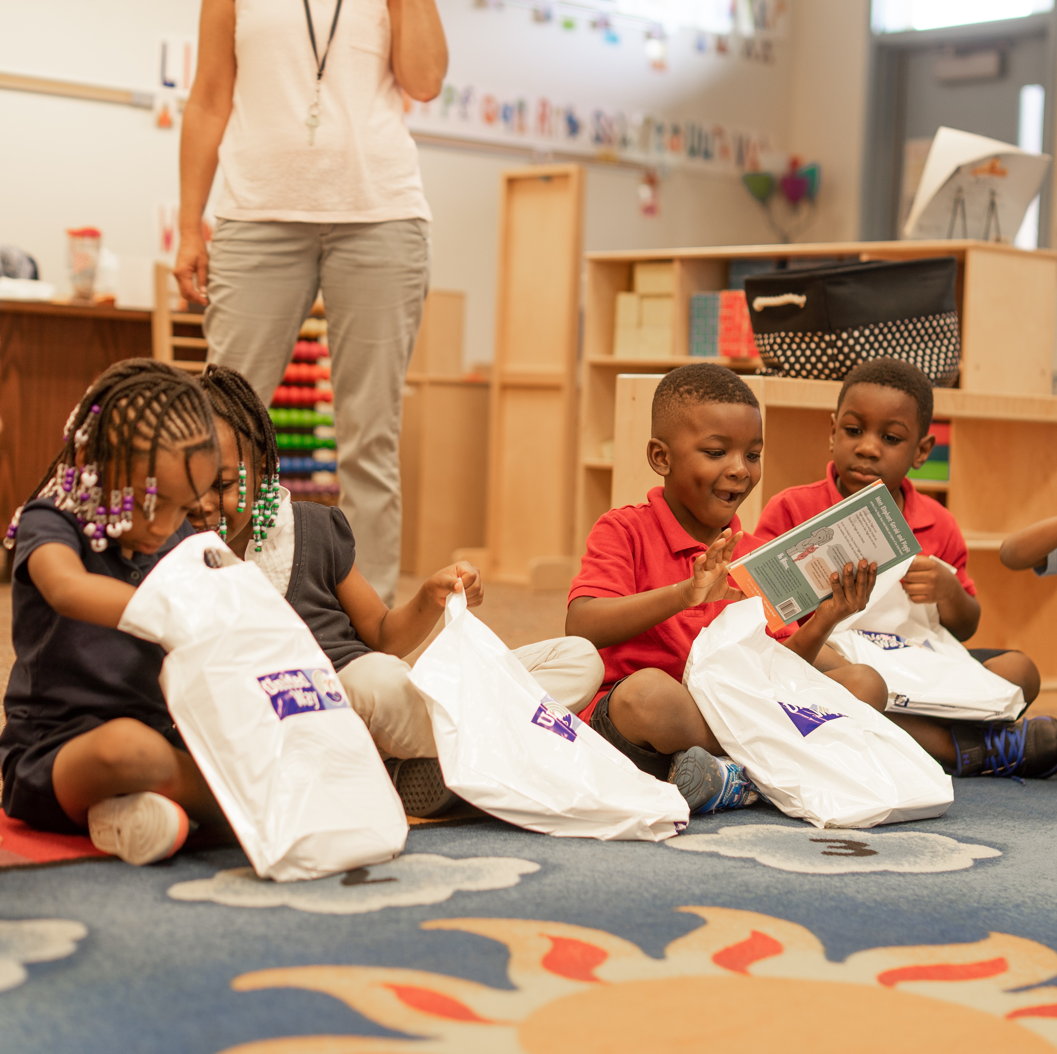 "Four children sit on a classroom rug and excitedly open their own small bag of children's books labeled with the Trident United Way logo."