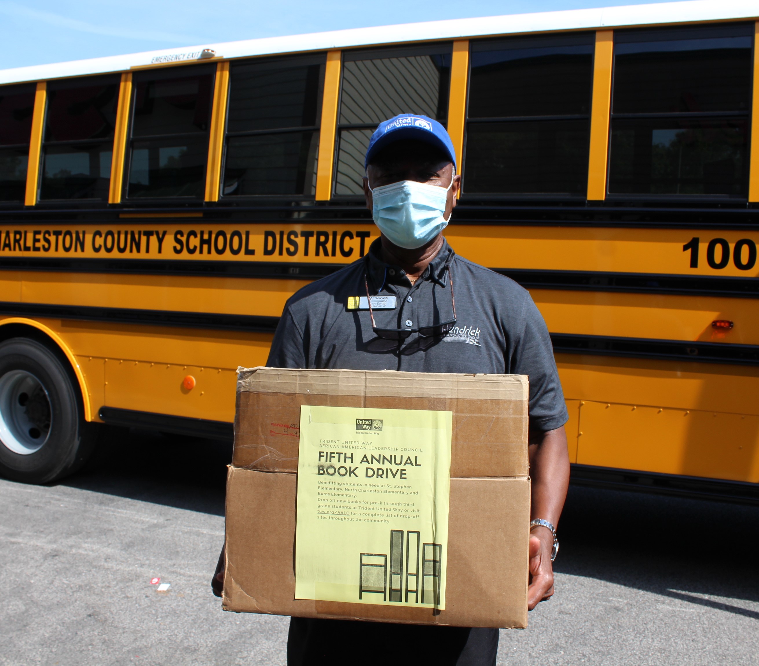 "AALC Member Don Smith stands in a mask in front of a school bus. He holds up a box labeled 'Fifth Annual Book Drive.'"