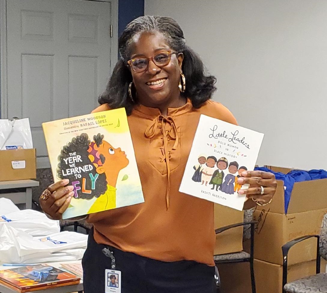 "AALC's Monifa Ellington smiles and holds up two children's books: 'The Year We Learned to Fly' with cover art of a young black girl, and 'Little Leaders: Bold Women in Black History' with a cover illustration of five black women."