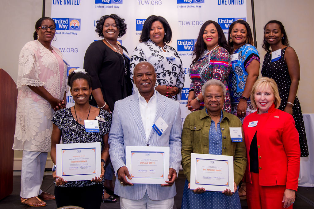 "A group of important people to the event stand and smile in front of a printed Trident United Way background. Three people sitting in front hold up award certificates."