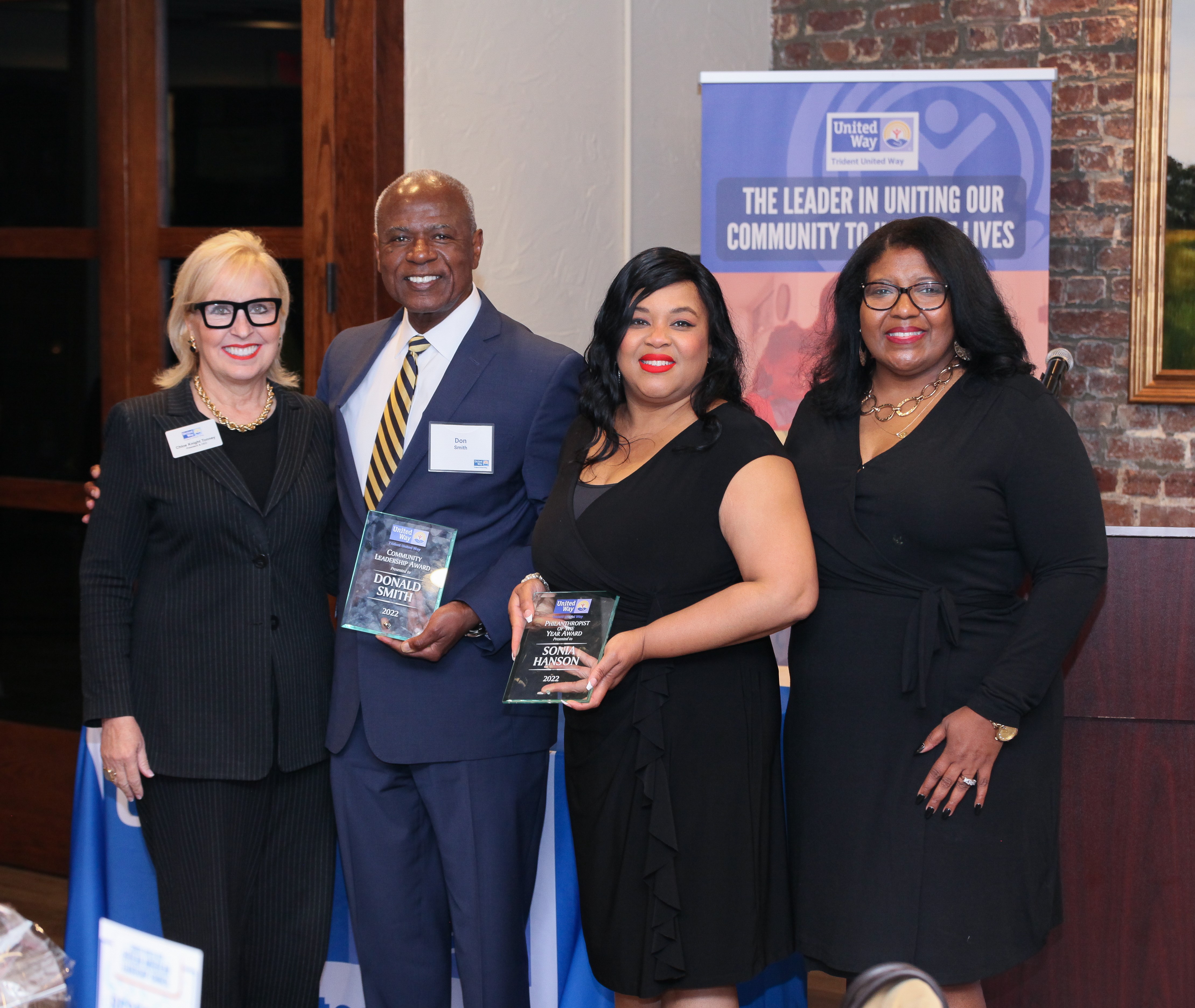 "Four people stand and smile for a picture. The two in the middle are award recipients Don Smith and Sonia Harrison holding up their plaques."