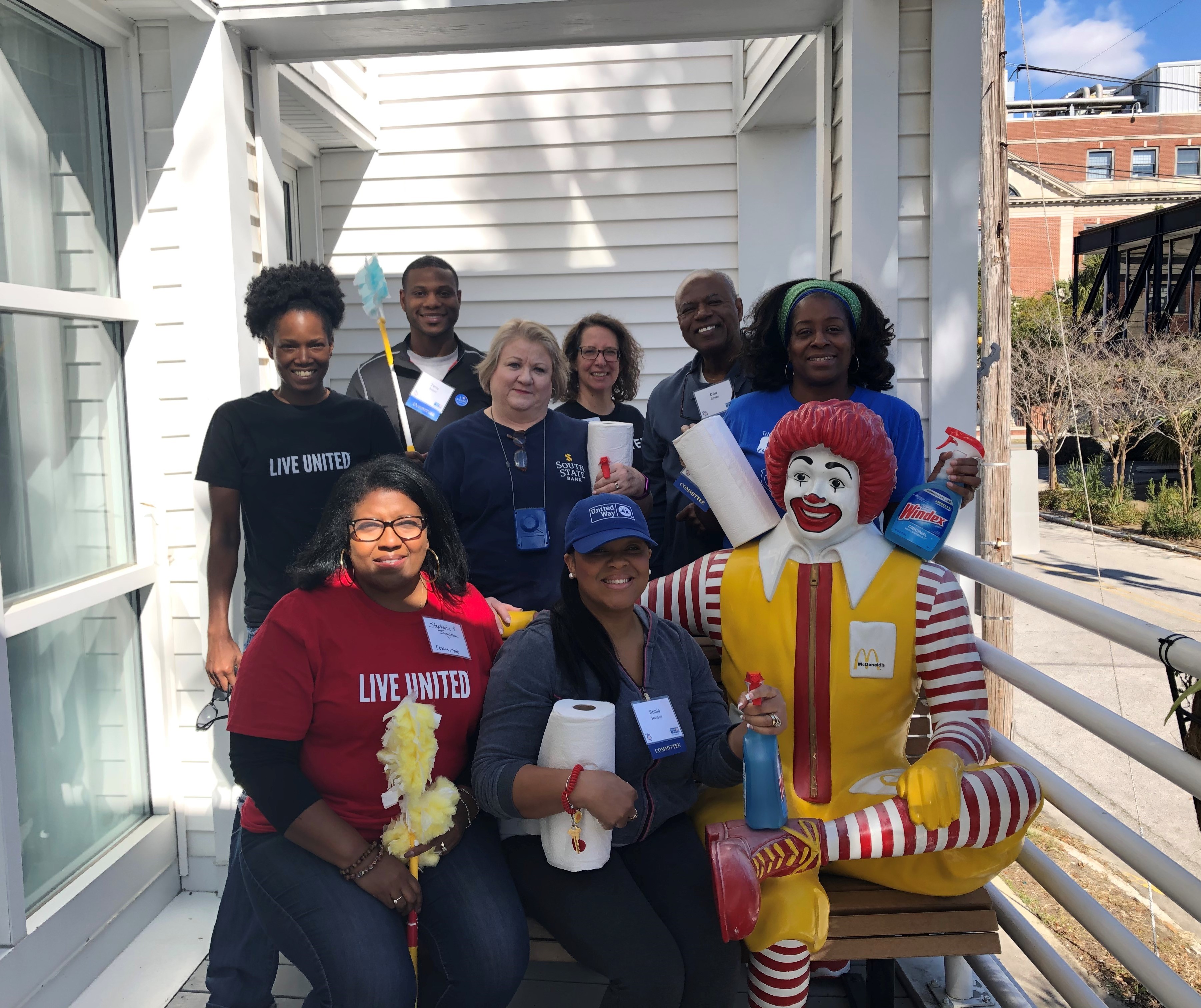 "A group of volunteers stands behind a bench smiling and holding cleaning supplies. Two volunteers sit on the bench next to a life-size statue that sits on the bench of character Ronald McDonald." 