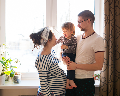 A pregnant woman smiles and looks at a man holding a toddler. They are standing in front of a window.