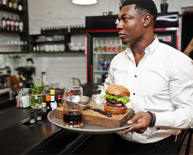 Black waiter holding a tray with an alcohol drink and burger in a nice restaurant. He's looking away.