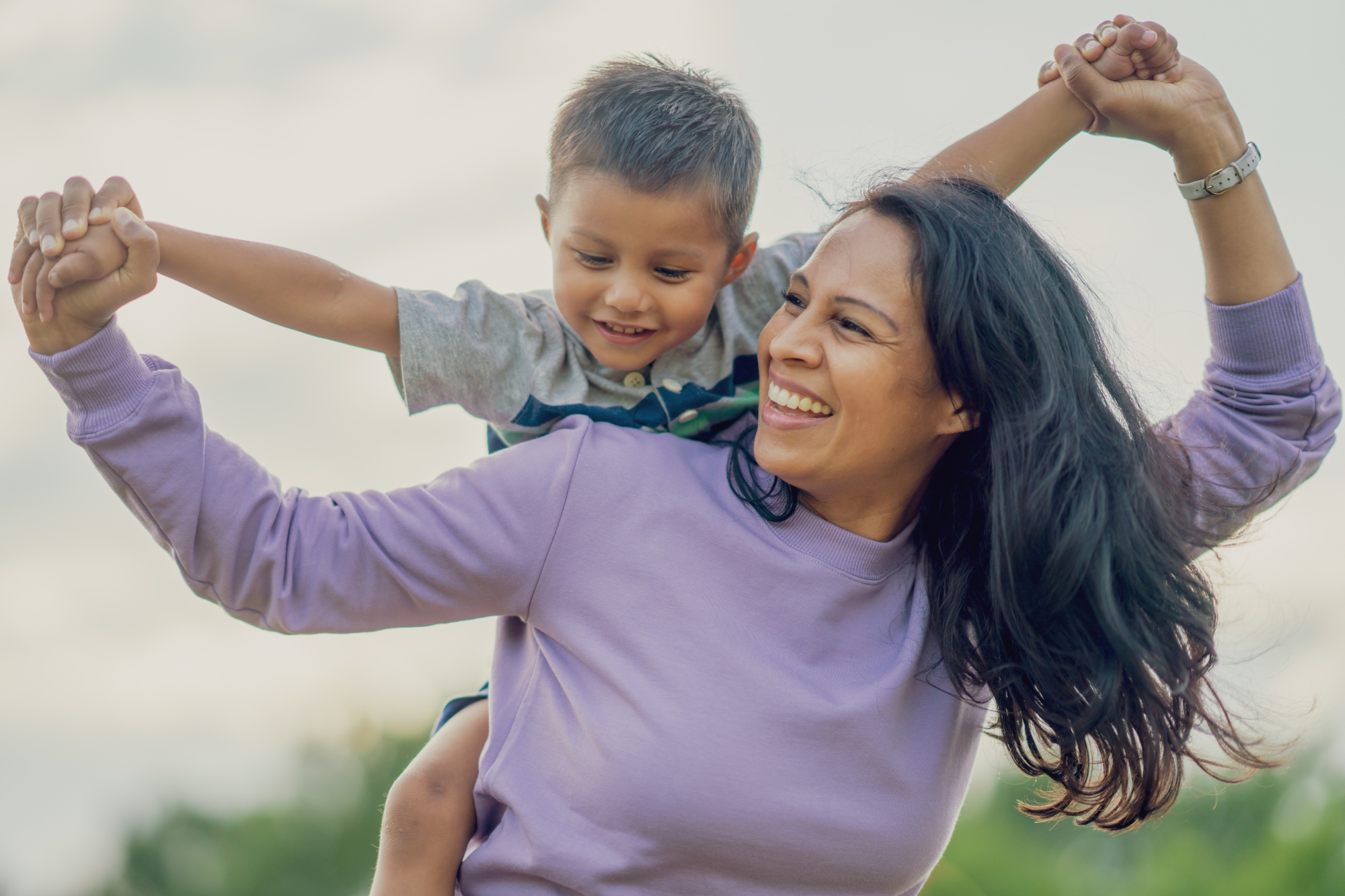 A young boy sits on the back of a woman, she is smiling and looking back at the boy, they are holding hands up in the air