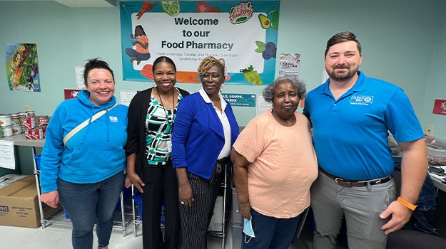 Photo of five individuals standing in the St. James Health and Wellness Clinic Food Pharmacy. An individual from the Lowcountry Food Bank is on the left, wearing a LCFB hoodie sweatshirt. The three individuals in the middle are from St. James Health and Wellness clinic. Two are wearing business casual attire, the volunteer is wearing casual attire. The final person in the photo on the right is from Trident United Way. He is wearing a blue polo with a TUW logo.