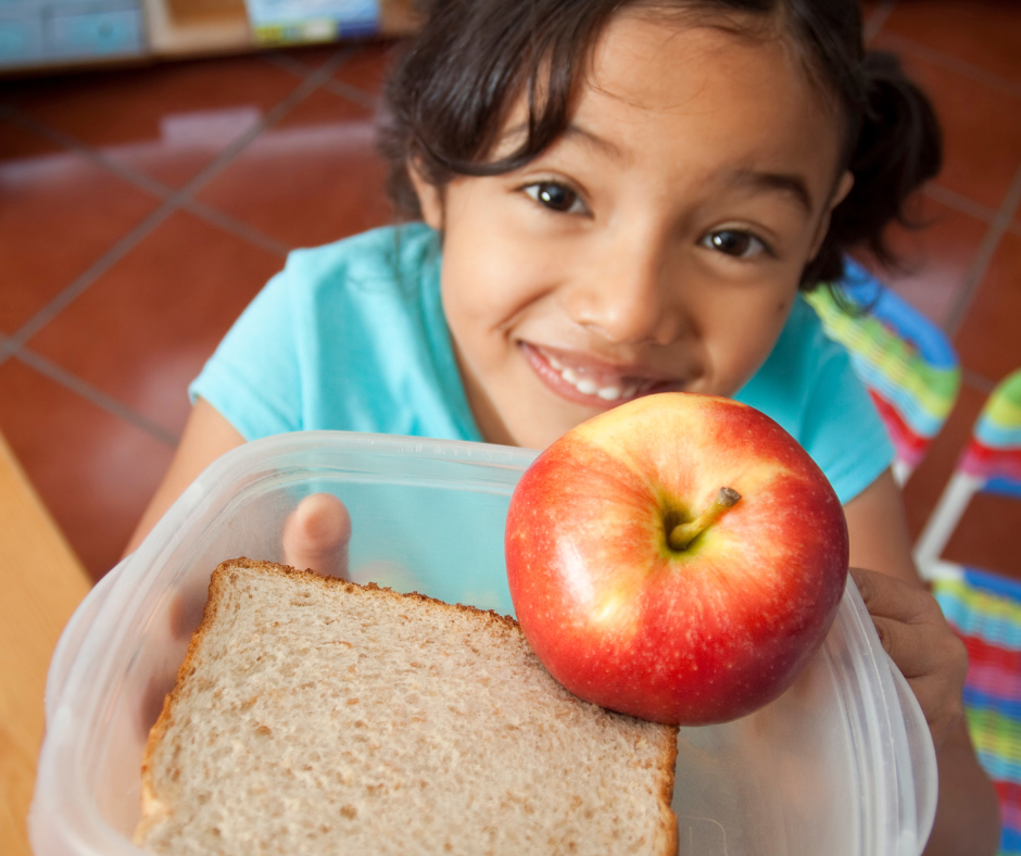 Young girl holding a tupperware container with a sandwich and an apple. She is smiling and is in a school.
