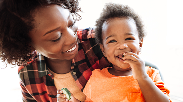 A mother holding her young child in her lap. The mother is smiling and looking at the child smiling,  child has a cucumber slice in both hands, placing one in her mouth