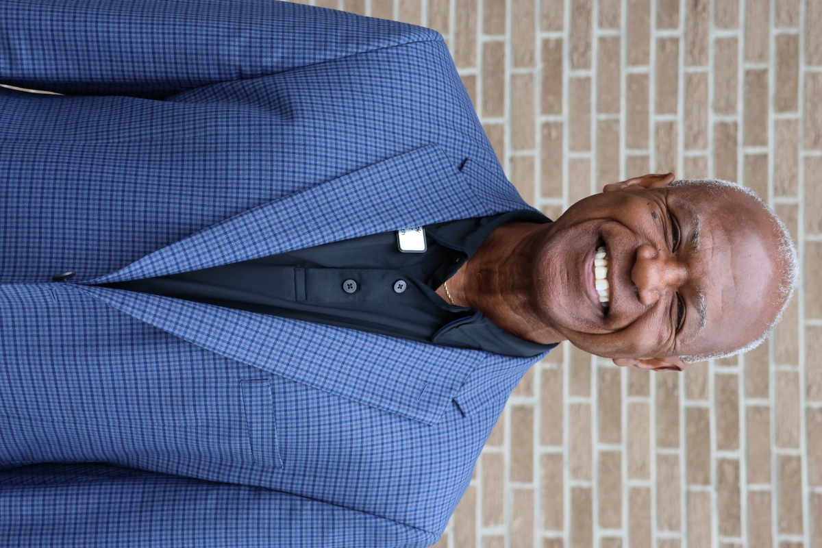 Don Smith standing in front a brick wall wearing a blue blazer and black polo shirt, smiling at the camera
