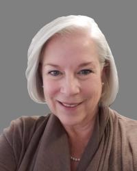 Cathy is posing in front of a grey background for her headshot. She is smiling and wearing a brown sweater.