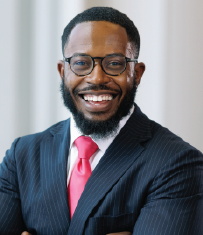 Headshot of Mike Sam. He is smiling and in a suit standing in front of a grey wall.