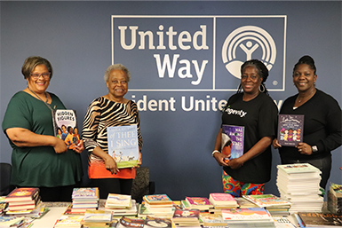 Four women standing in front of the United Way logo behind a table of books, each with one in their hands at the book drive