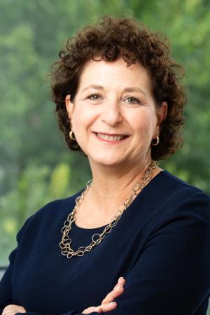 Photo of Cindy Brams. She has short, dark curly hair, wears small gold hoops and a gold necklace. She is wearing a dark blue top and is smiling at the camera.