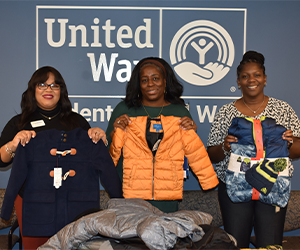 Three members of the AALC holding up donated coats in front of a Trident United Way logo