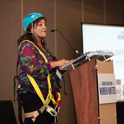 Teresa Vaughn speaking on stage wearing rapeling gear from Over the Edge