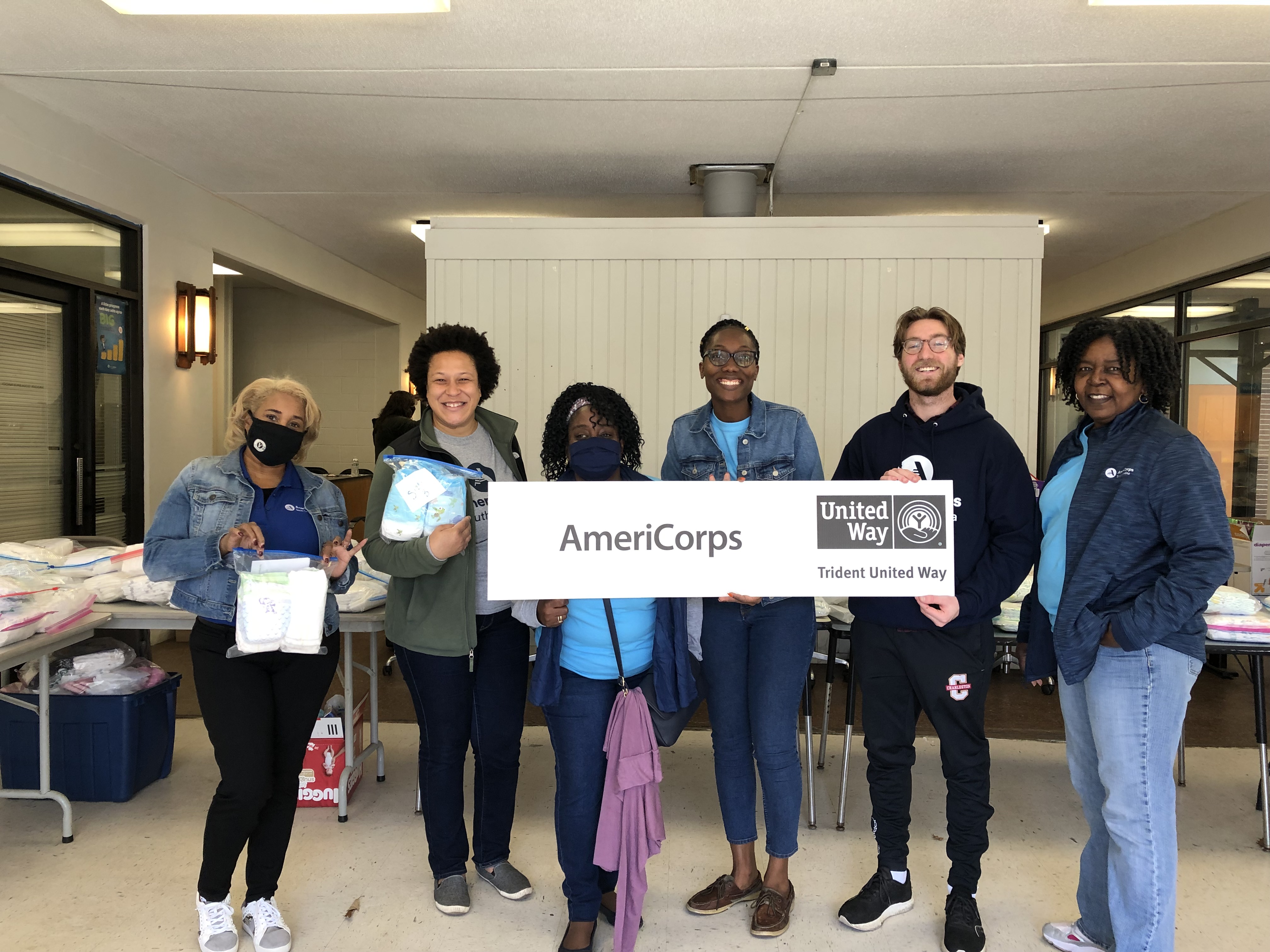 Six AmeriCorps members holding a sign that says AmeriCorps, holding ziploc bags filled with diapers from volunteer activity