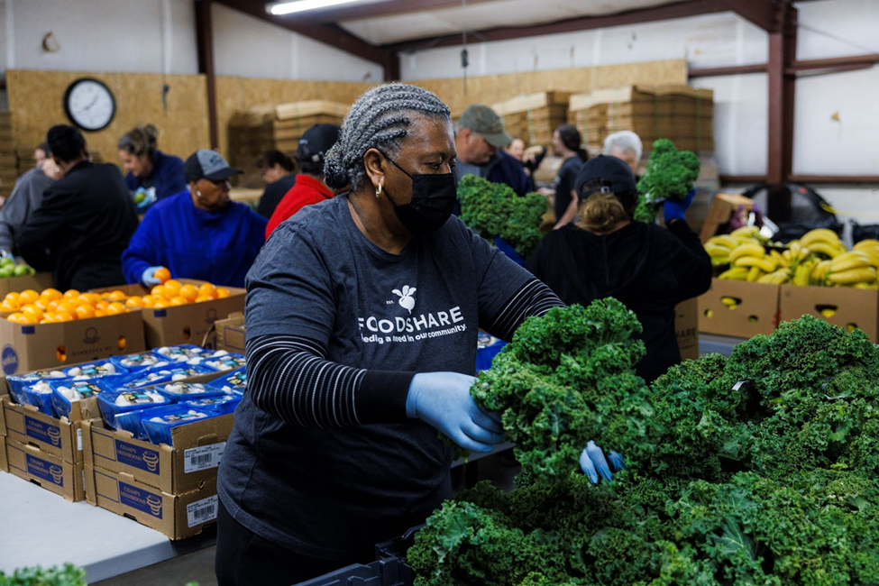 Older, African American woman is picking a bushell of kale out of a pile of kale. She has on a black face mask and blue kitchen gloves. She is wearing a black t-shirt with the foodshare logo over a longsleeve striped shirt