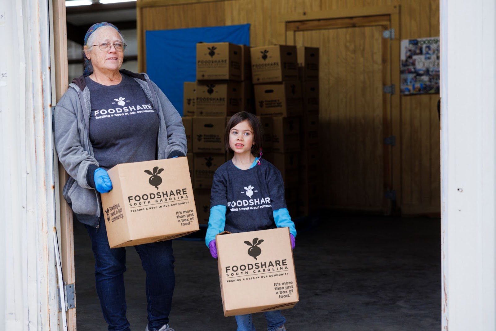 An older white woman and younger white girl stand holing FoodShare South Carolina boxes. The older white woman has a scarf on her head and is wearing a grey FoodShare SC shirt under a gray zip up hoodie. The girl is wearing a grey FoodShare SC shirt over a bright blue long sleeve shirt. They are standing in a large doorway with FoodShare boxes behind them.