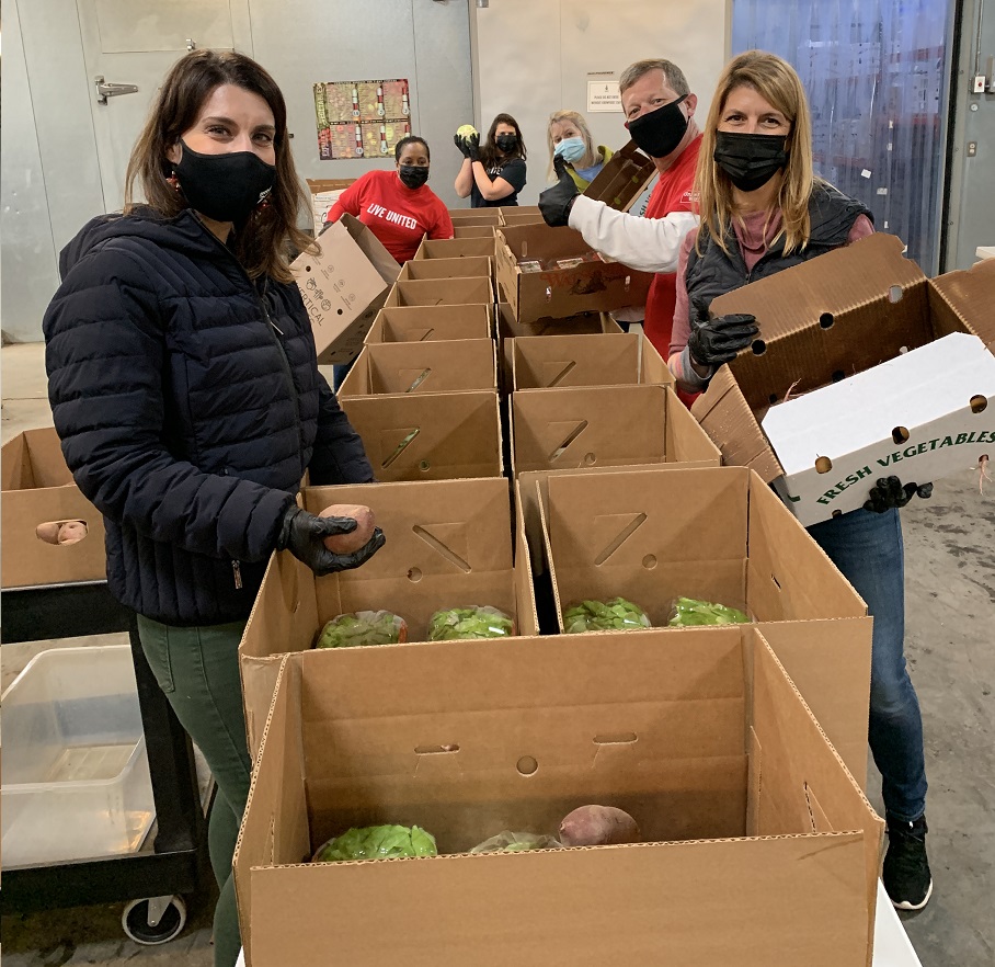 Trident United Way staff members pose for photo while volunteering to pack food boxes. Two staff volunteers hold produce in their hands, one holds a box. They are in a warehouse setting
