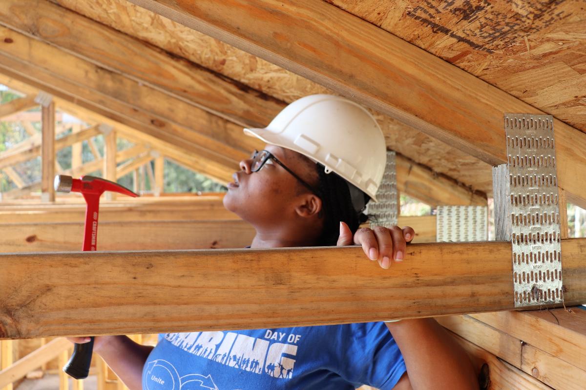 Volunteer holding a hammer, wearing a hardhat and a Day of Caring shirt helping to build the frame of a house for Habitat for Humanity