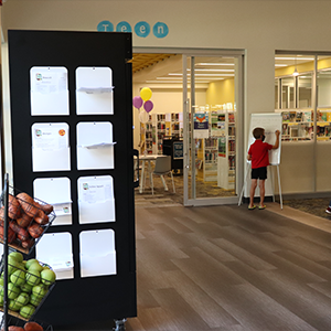 a photo showing the side view of a fridge with manilla folders containing recipe cards, the three tiered stand containing produce baskets. In the background of the photo is a section of the library