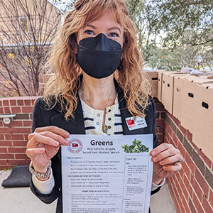 Rep from Lowcountry Food Bank holding a description flyer for Greens, standing in front of the large cardboard boxes containing produce