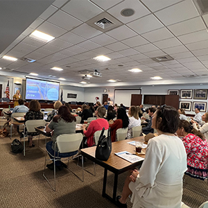 Several individuals seated classroom style in a large room in an office building in a presentation