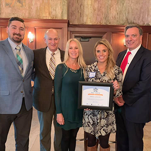 Five individuals wear professional clothes posing for photo, one holds a certificate from the Healthy Business Challenge