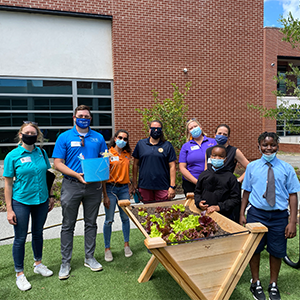 A group of teachers and students stands behind a garden bed with greenery, a Trident United Way staffer holds a blue bucket containing produce