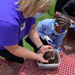 A teacher holds dirt in her hands above a small bucket containing dirt while a young girl looks on