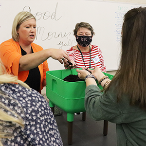 Three indivduals are sticking their hands in a large bucket of dirt during an instructional session for teachers