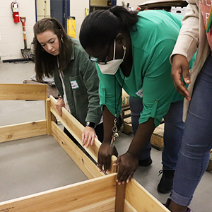 Two individuals are helping to put a plank of wood in place to build garden beds in a large warehouse type room