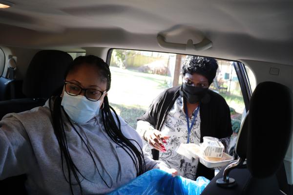 AmeriCorps member Shawana Paige sits in a car holding a covered hot meal tray while Brenda Bunch from Dorchester Seniors stands outside the car handing a tray to Shawana.