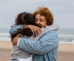 two women hugging on a beach. One is wearing a jean jacket the other a grey sweatshirt. It looks like a cold and cloudy day.