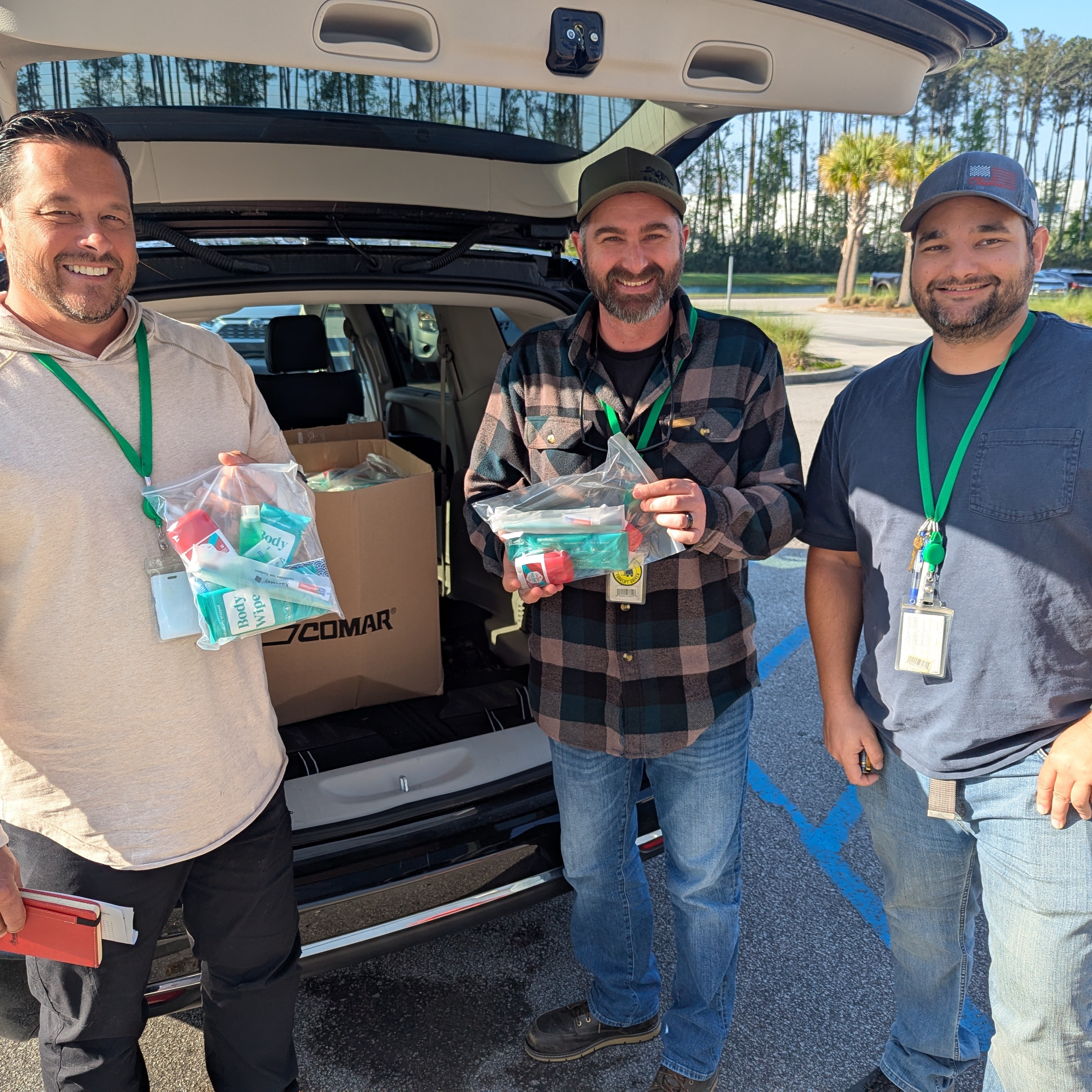 3 participants in Day of Action stand behind a car's full trunk while holding additional wellness products they packed in kits.