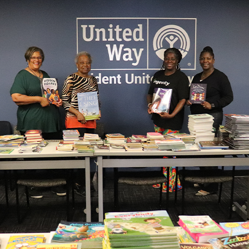 Four members of the African American Leadership Council stand in front of a wall with a Trident United Way logo on it. The four members are holding a book in their hands, smiling and facing the camera. In the foreground of the photo are stacks of books lined up on tables