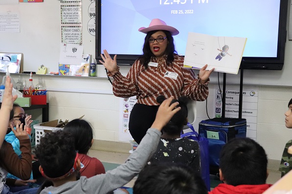 Photo of volunteer Sonia Hanson reading to a classroom. She wears a pink cowboy hat and has her arms up while holding a book so students can see illustrations. Student visible in photo are mimicking how she is holding up her arms.