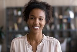 Photo of Black woman looking at camera and smiling. She is inside a book shop, and wearing a light pink top. She has short, curly hair.