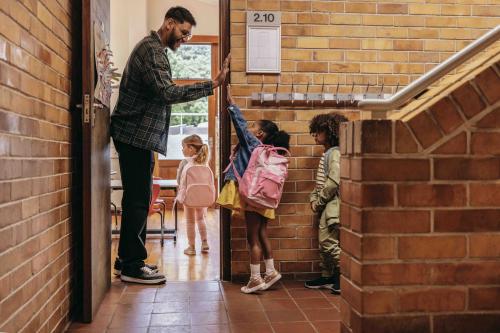 A man is giving a high five to a young student in a school hallway outside of a classroom.