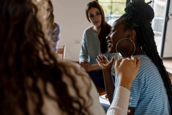 A group of women sit in a circle, the focus is on a younger woman who is speaking and smiling. A hand sits on her shoulder to in a comforting way.