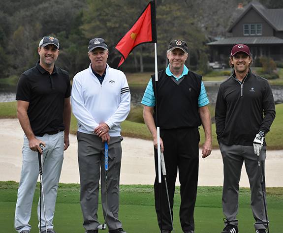 "Four men stand and smile holding golf clubs. Behind them is a bit of golf course and a small pond, a flag, and a house."
