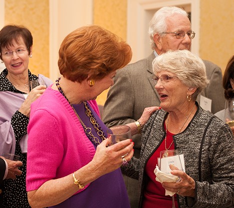 "A woman facing away smiles and rests her hand on another woman's shoulder. The woman to the right holds a drink and speaks. The room in the background has a subtle gold wallpaper."