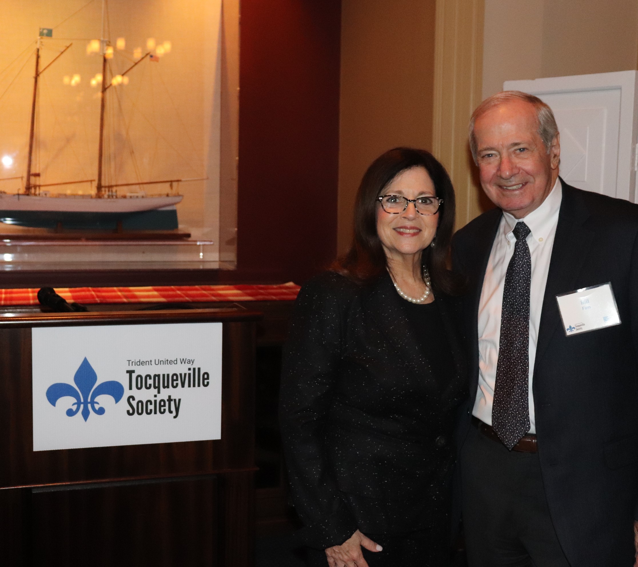 "On the left, a model ship rests on a table behind a window, behind another table with a sign reading "Trident United Way Tocqueville Society. On the right, co-chairs Anita Zucker and Bill Finn stand and smile."