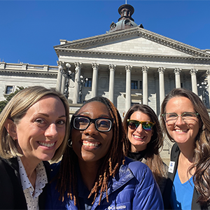 Four adult women are standing outside of the SC State House, smiling at the camera.